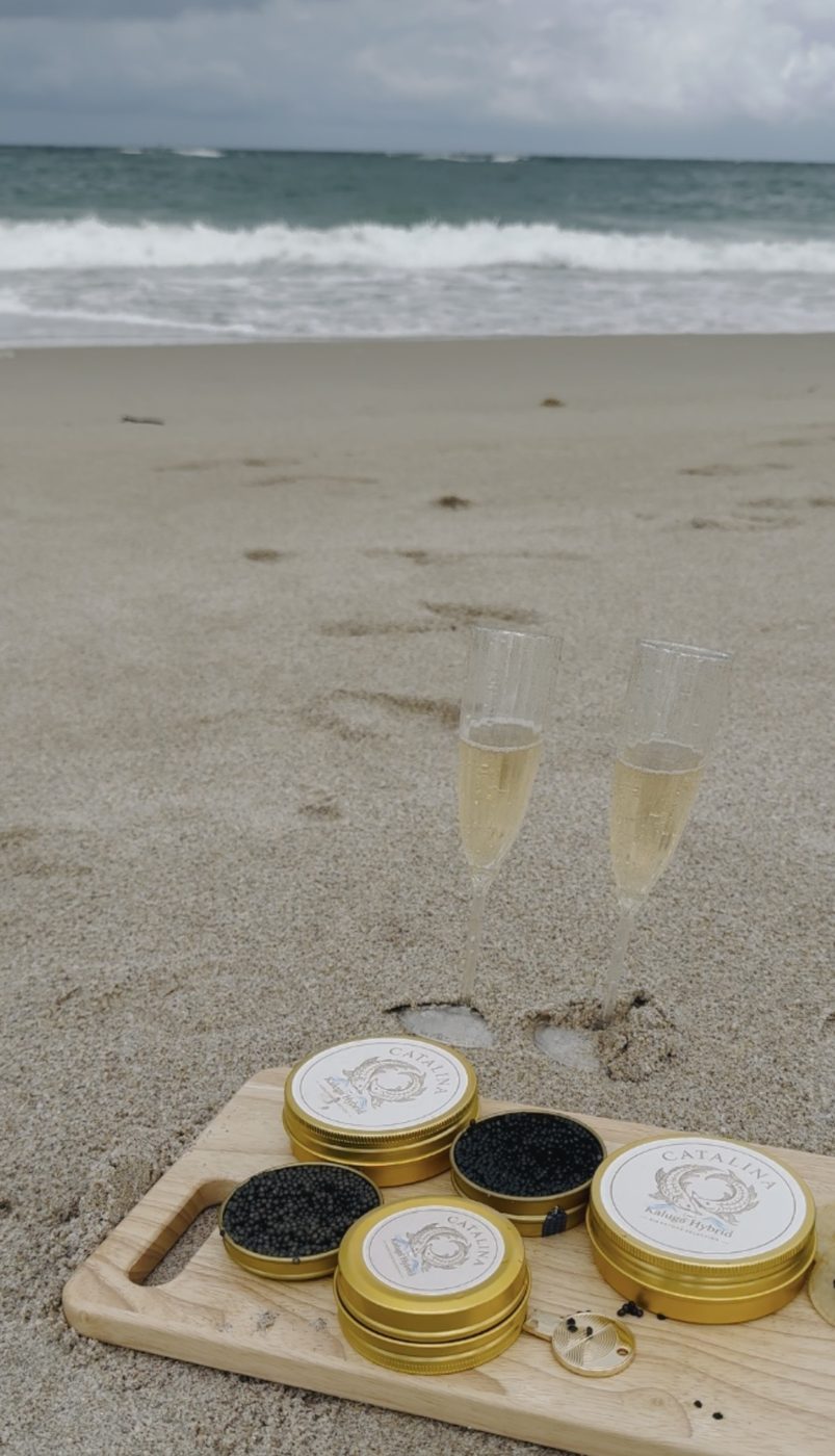 A board of gold tins and two champagne flutes in the sand by the surf