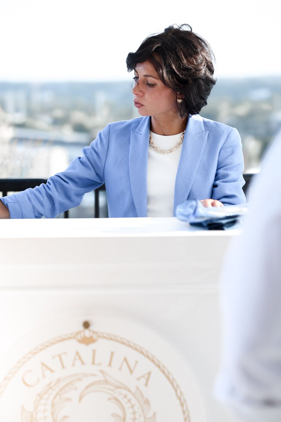 A Catalina host in a pale blue blazer arranging navy napkins at the crested caviar cart, city skyline behind