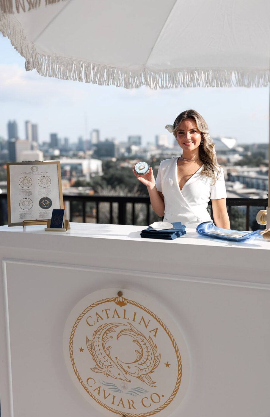 A Catalina host in white at the crested caviar cart under a fringed umbrella, city skyline behind