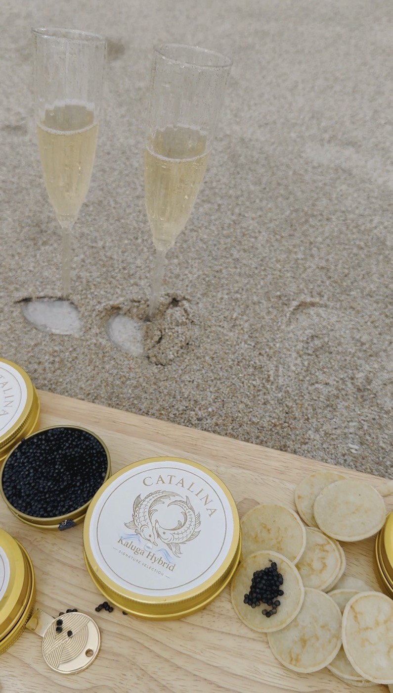 Gold Catalina caviar tins and blinis on a board with two champagne flutes standing in the sand